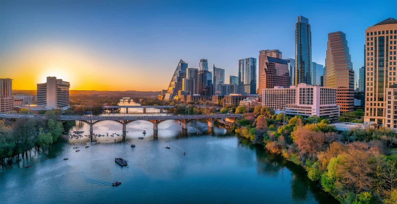 Austin, Texas- Cityscape with Colorado River in the middle against the sunset sky. There are boats near under the bridge at the front and a view of skycrapers on the side with reflective glass walls.